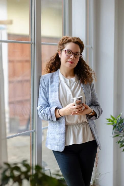 Várnai-Látos Szilvia life és mindfulness coach portré – az ablaknál állva egy bögre kávét tart a kezében.