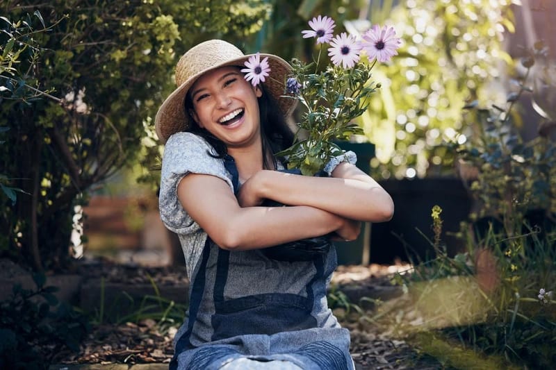 Tudatos jelenlét mindfulness tanácsadással - a kertben egy mosolygós kertész lány kezében lila kerti virágokkal, fején kalappal.