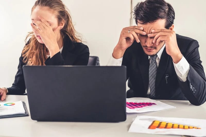 Kiégés feldolgozása coachinggal - Manager nő és férfi az asztalnál ülve, fogják a fejüket a fáradtságtól, laptop és grafikonokat ábrázoló papírok előttük.