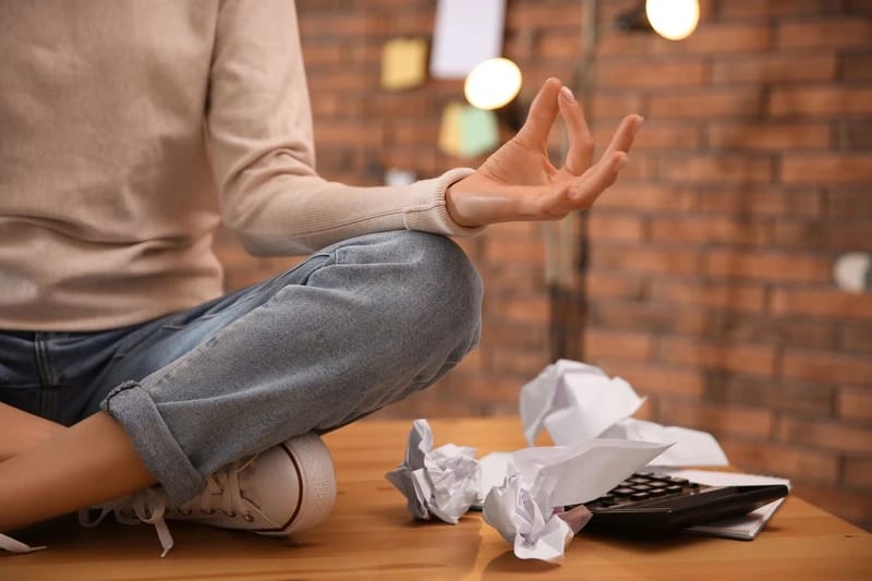 Stressz oldása coaching tanácsadással - egy irodai asztalon meditatív ülésben és kéztartással ülő lány körülötte összegyűrt papírokkal és számológéppel.