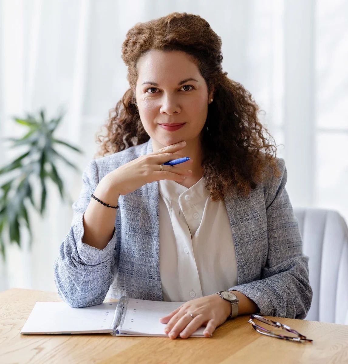 Várnai-Látos Szilvia life és mindfulness coach portré – az asztalnál ülve tollat tartva a kezében néz előre, egy jegyzetfüzet és szemüveg előtte.