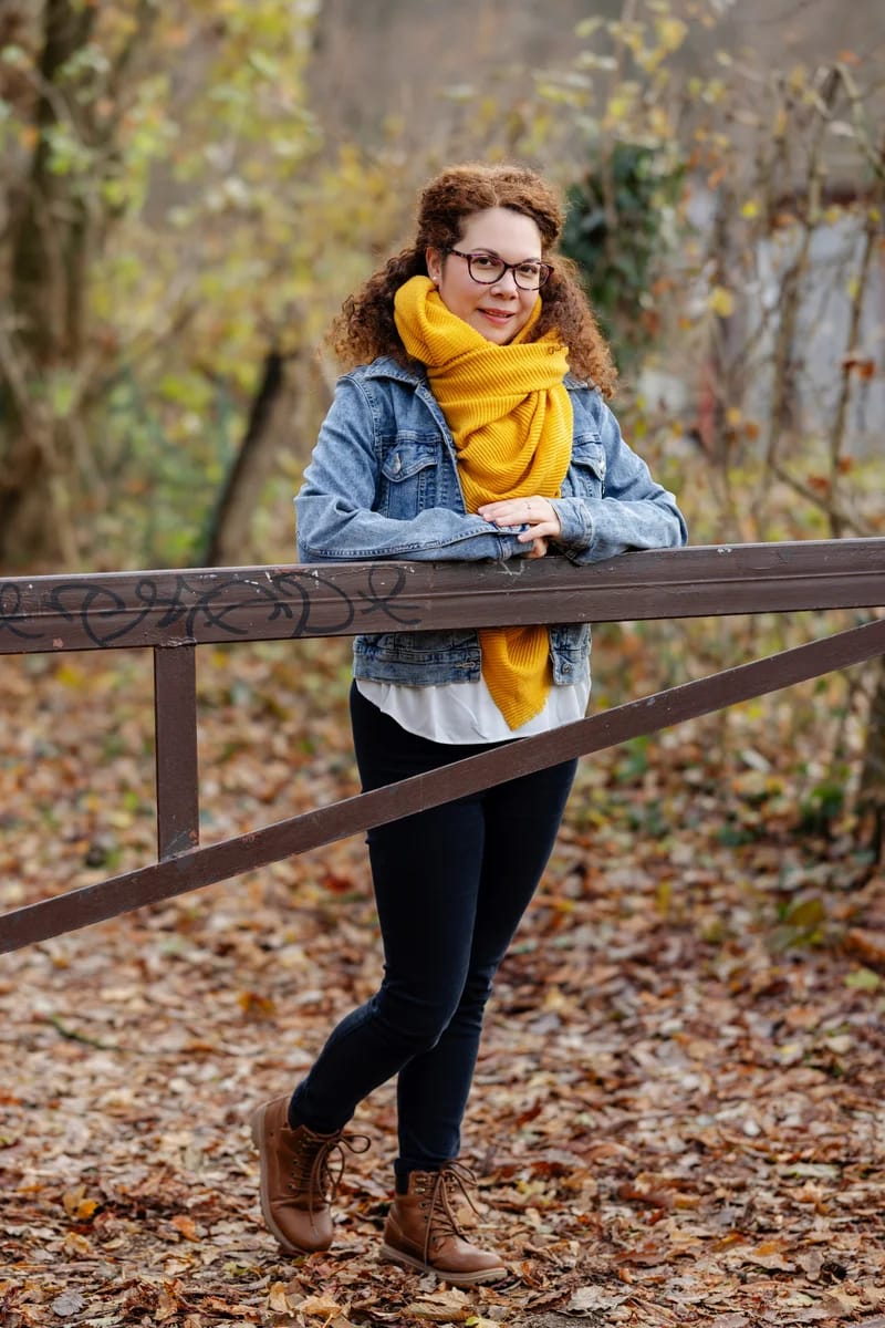 Várnai-Látos Szilvia – mindfulness séta az őszi hangulatú erdőben. Szilvi egy kapura könyököl, farmerkabát, sárga sál, fekete farmer és barna csizmát visel.